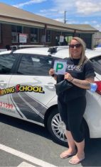 A new driver in sunglasses holding a green P plate beside a white Affordable Driving School car