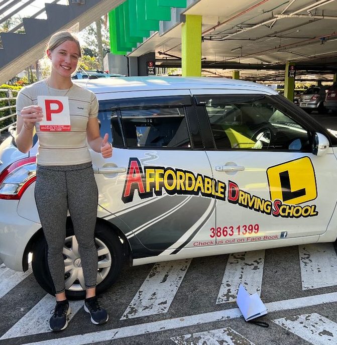 A new driver holding a P plate, thumbs up, beside a white Affordable Driving School car in a car park