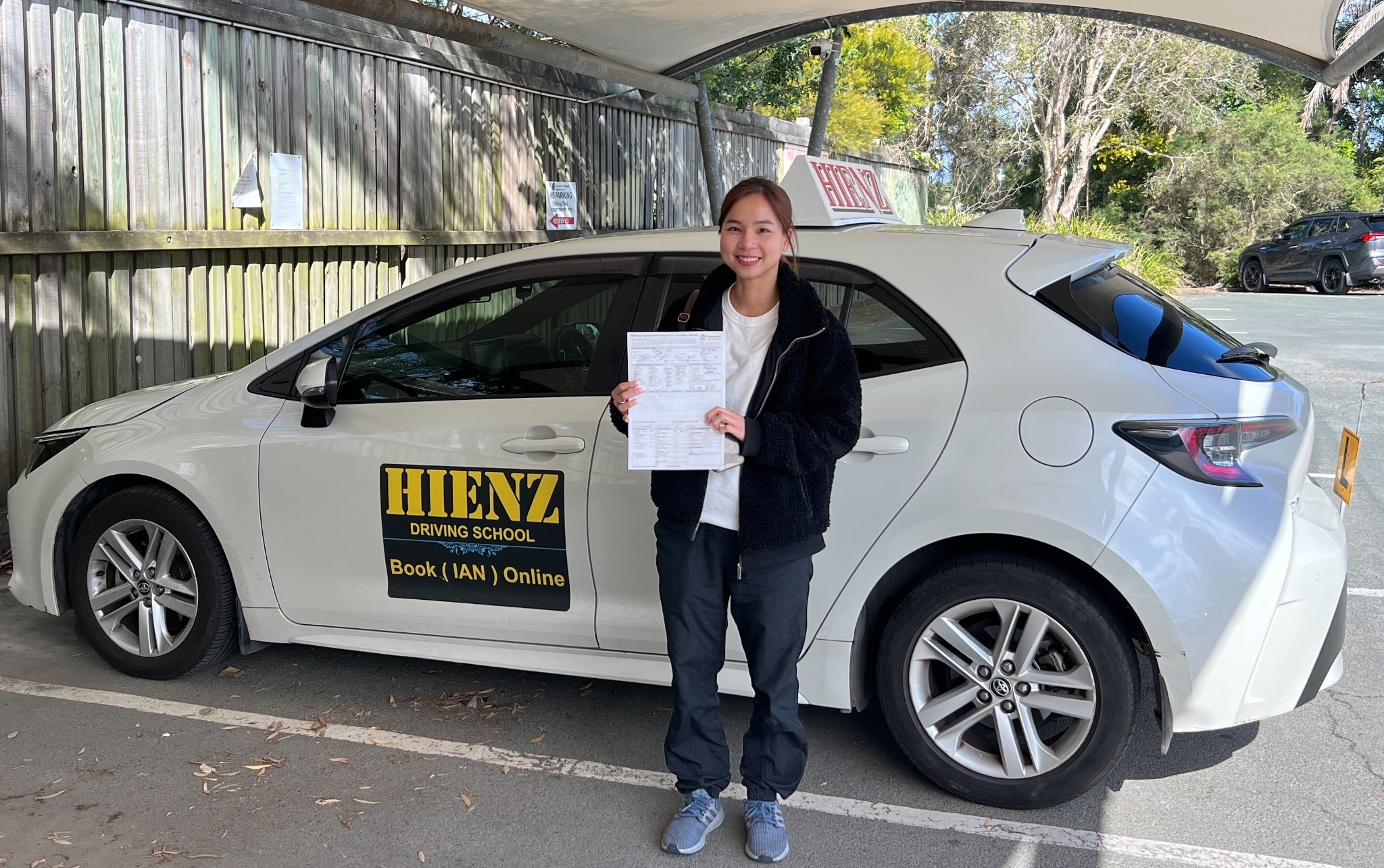 Successful HIENZ student holding driving test paperwork beside a white Toyota with HIENZ driving school signs.