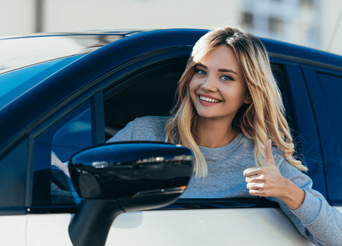 A smiling young woman making a thumbs-up gesture inside the car.