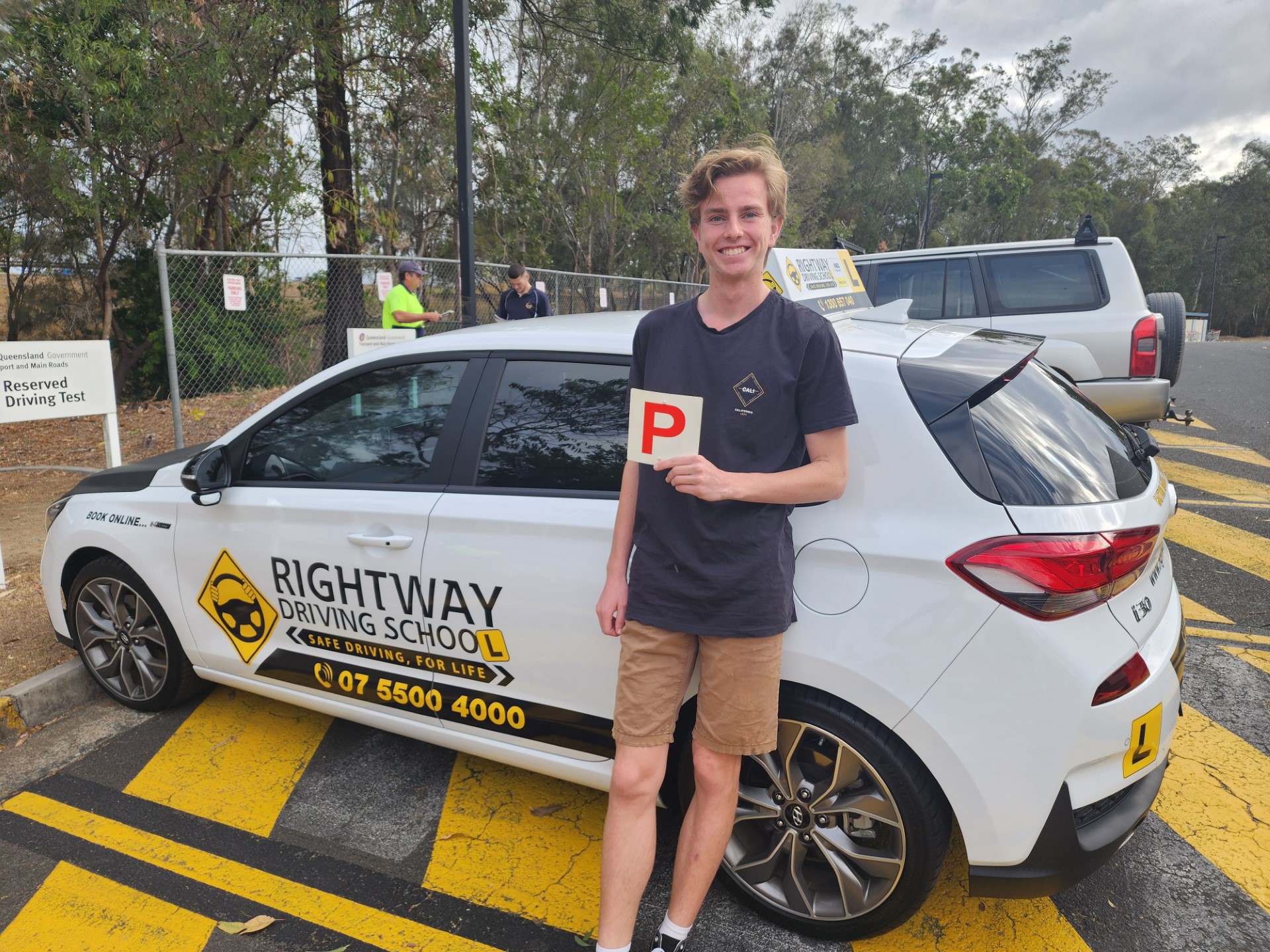 Person in a dark T-shirt and pale shorts holding a red P plate beside a white Rightway learner car, with trees behind.