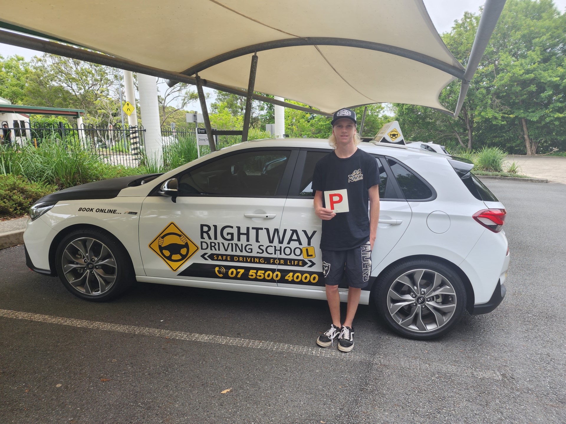Person in a dark cap and black top holding a red P plate beside a white Rightway learner car under a canopy.