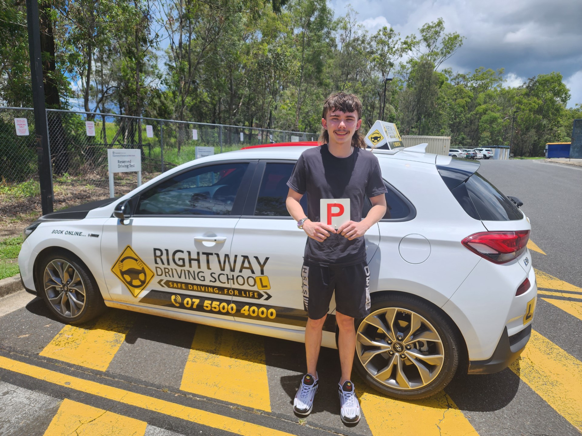 Person in a dark top and shorts holding a red P plate beside a white Rightway learner car, with trees behind.