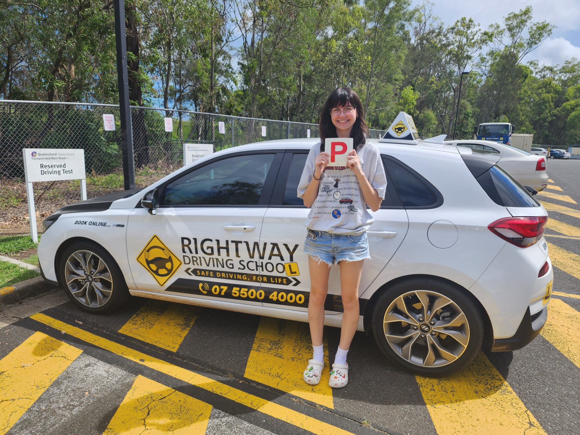 Person in a striped top holding a red P plate beside a white Rightway learner car, with trees behind.
