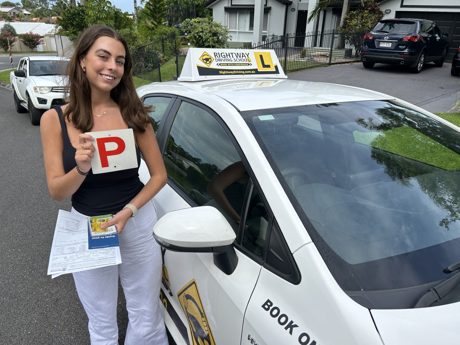 Person in a dark top and pale trousers holding a red P plate beside a white learner car with a Rightway roof sign.