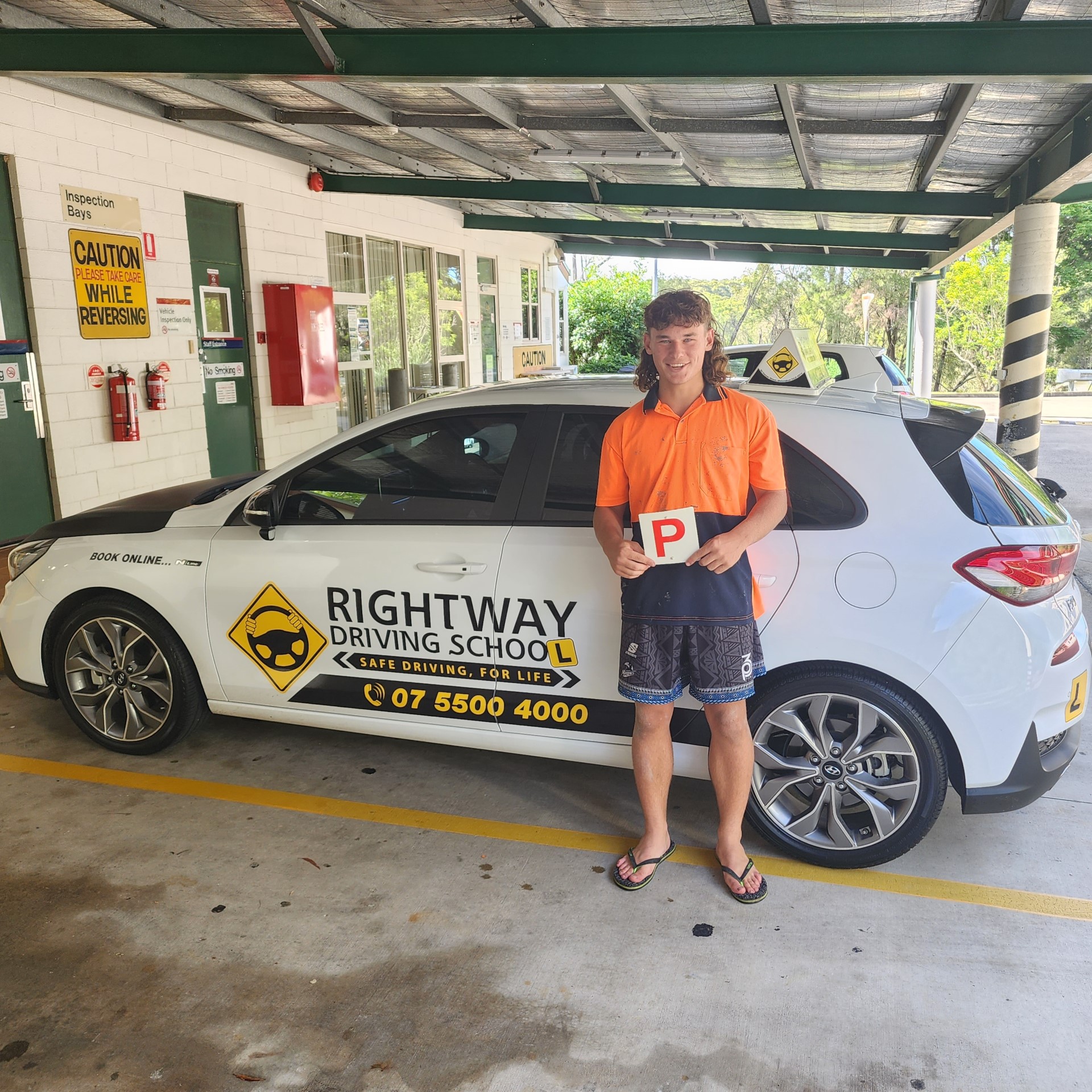 Person in an orange shirt holding a red P plate beside a white Rightway learner car under a roof.