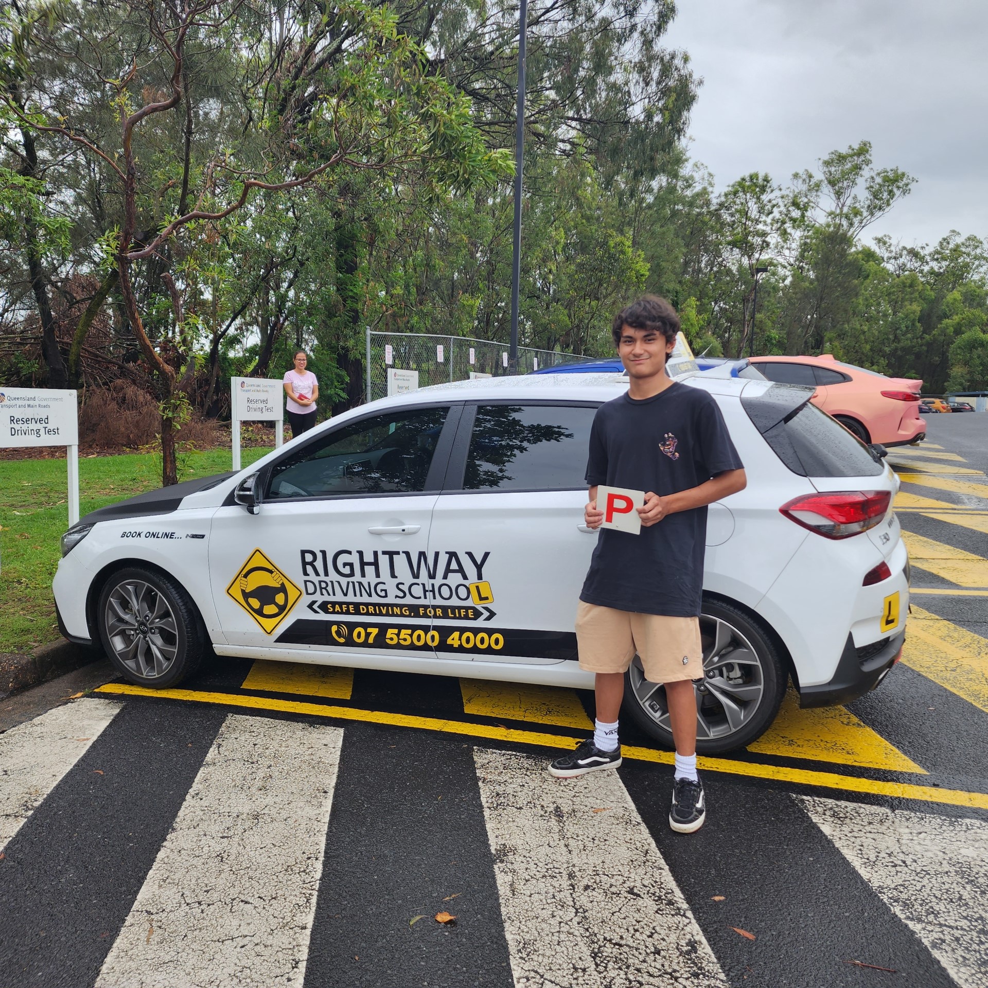 Person in a dark top and pale shorts holding a red P plate beside a white Rightway learner car outdoors.