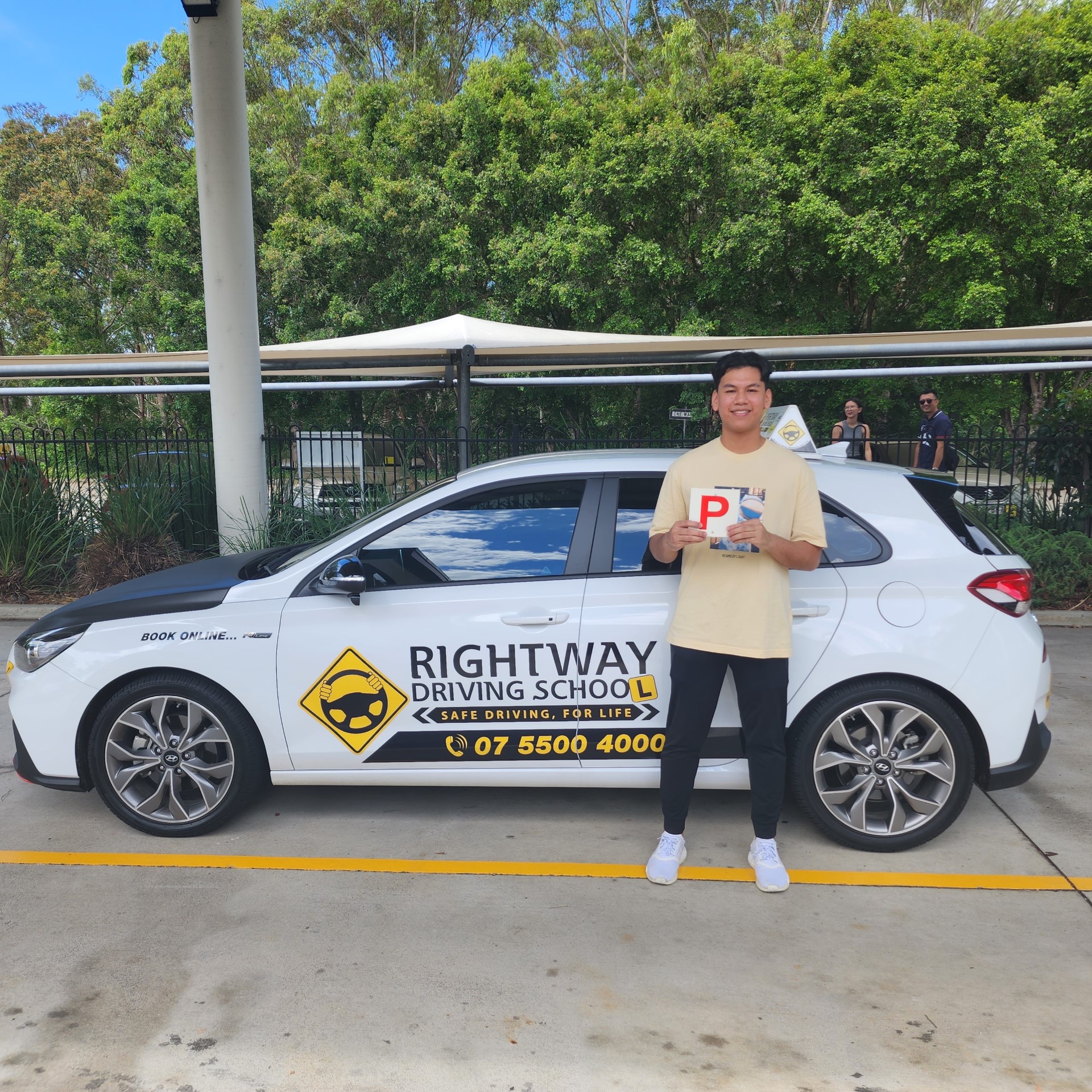 Person in a pale T-shirt holding a red P plate beside a white Rightway car photographed side-on with greenery behind.