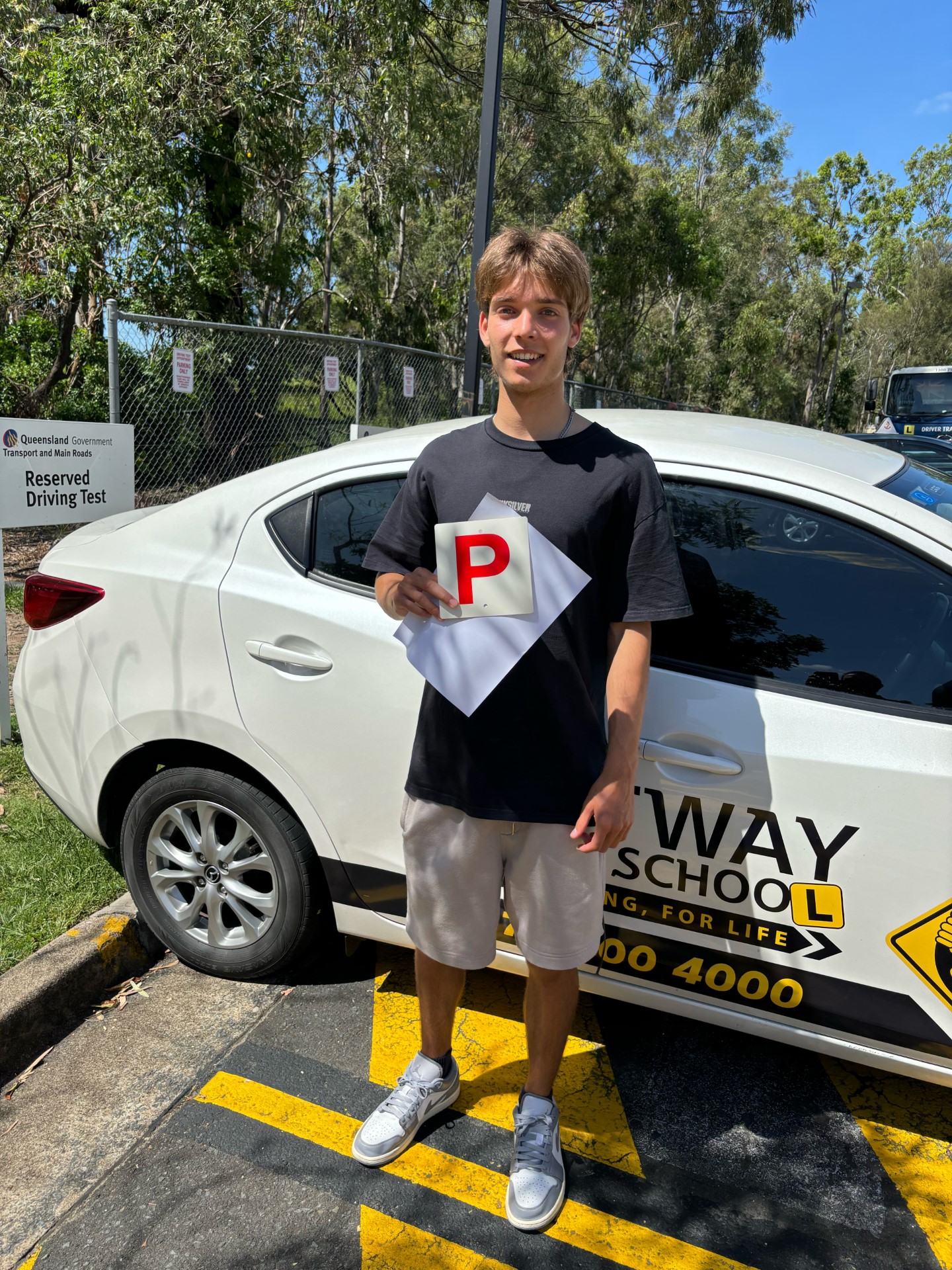 Person in a black T-shirt and pale shorts holding a red P plate beside a white Rightway learner car.