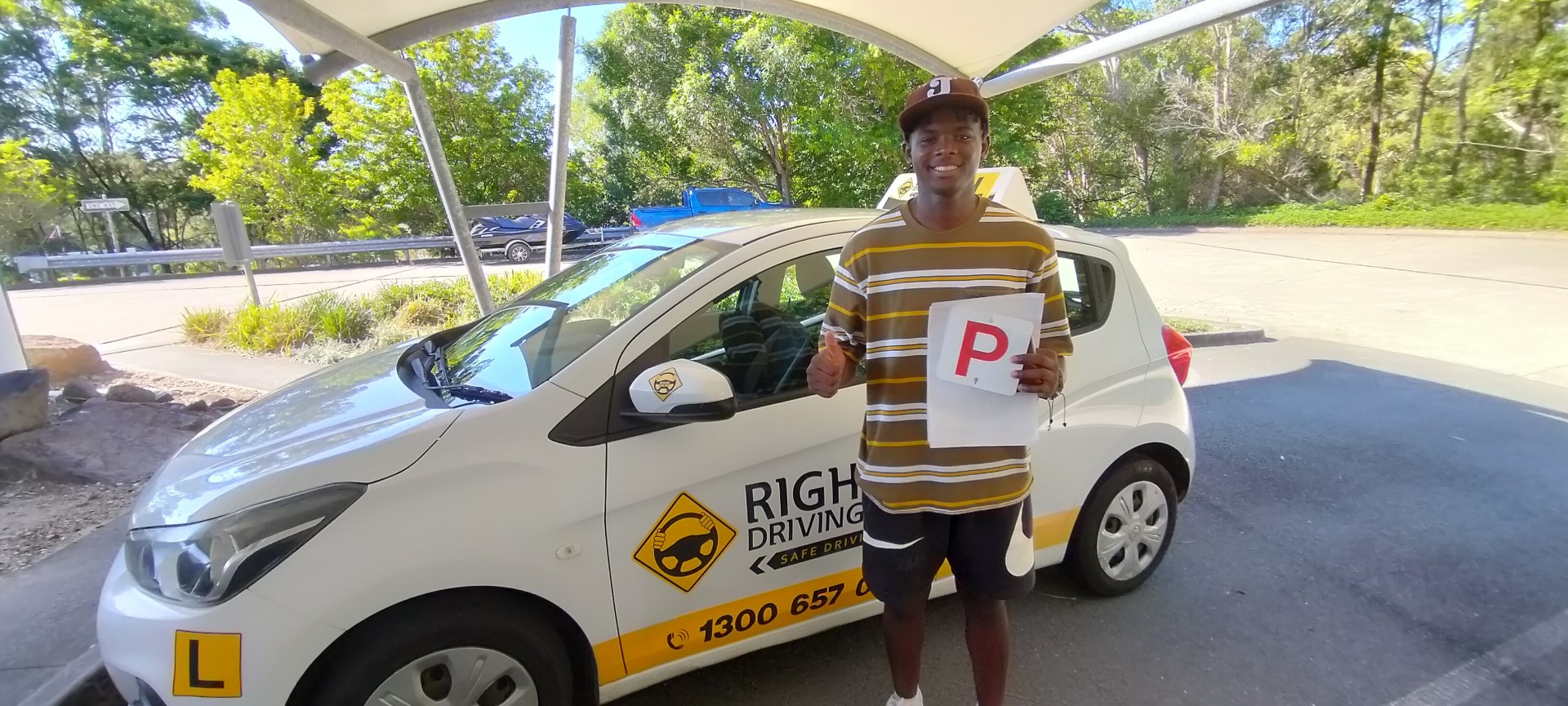Person in a striped shirt holding a red P plate beside a white Rightway learner car under a canopy.