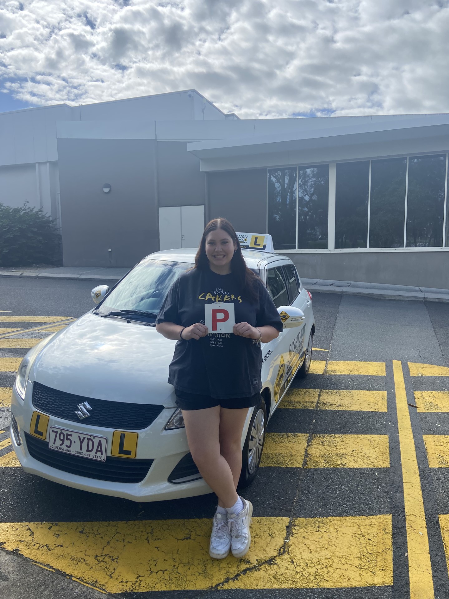 Person in a dark outfit holding a red P plate at the front of a learner car outside a building.