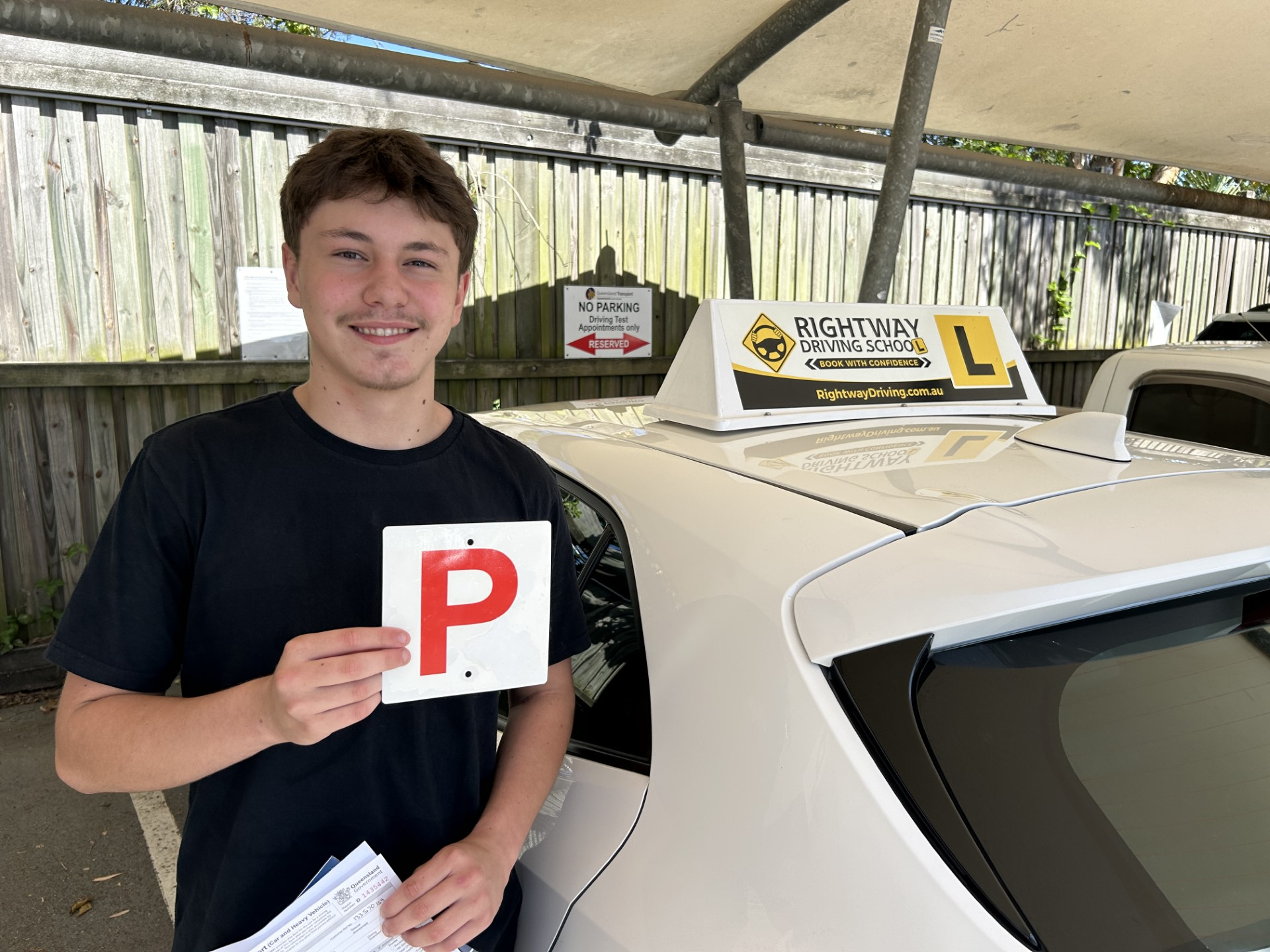 Person holding a red P plate beside a white Rightway learner car