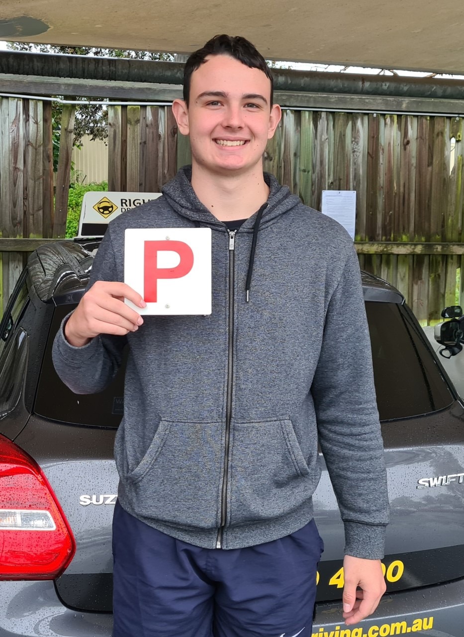 Person in a grey zip-up hoodie holding a red P plate beside the rear of a dark car.