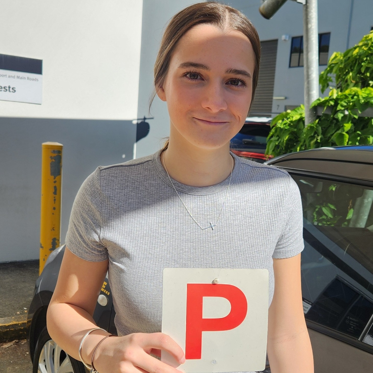 Person in a grey top holding a red P plate beside a dark car, with a building behind.
