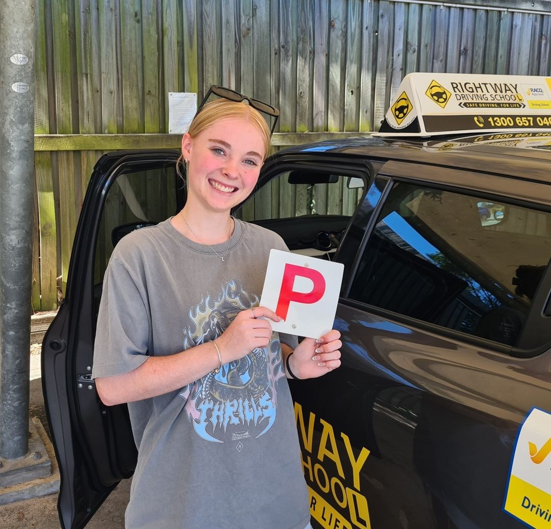 Person in a grey printed T-shirt holding a red P plate beside a dark car with a Rightway roof sign.
