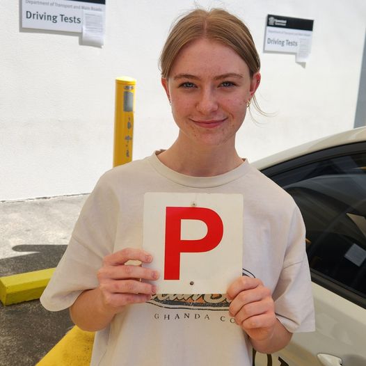 Person in a pale printed T-shirt holding a red P plate beside a car and building.