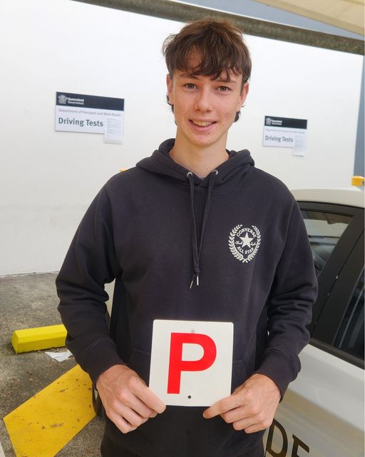Person in a dark hoodie holding a red P plate, with a car and driving-test sign in the background.