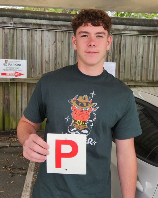 Person in a dark printed T-shirt holding a red P plate under a canopy beside a car.