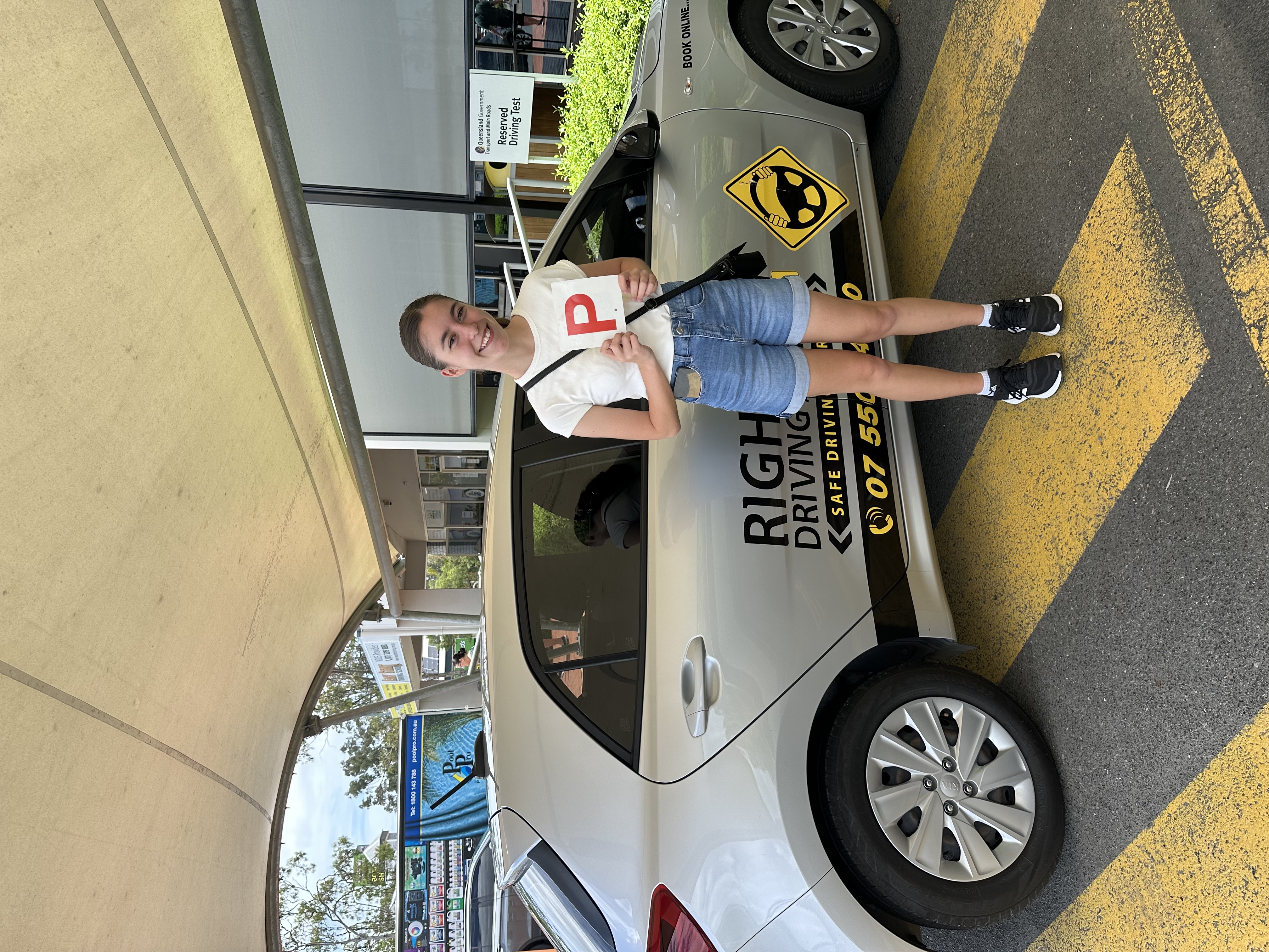 Person in a pale top and denim shorts holding a red P plate beside a silver Rightway car under a canopy.