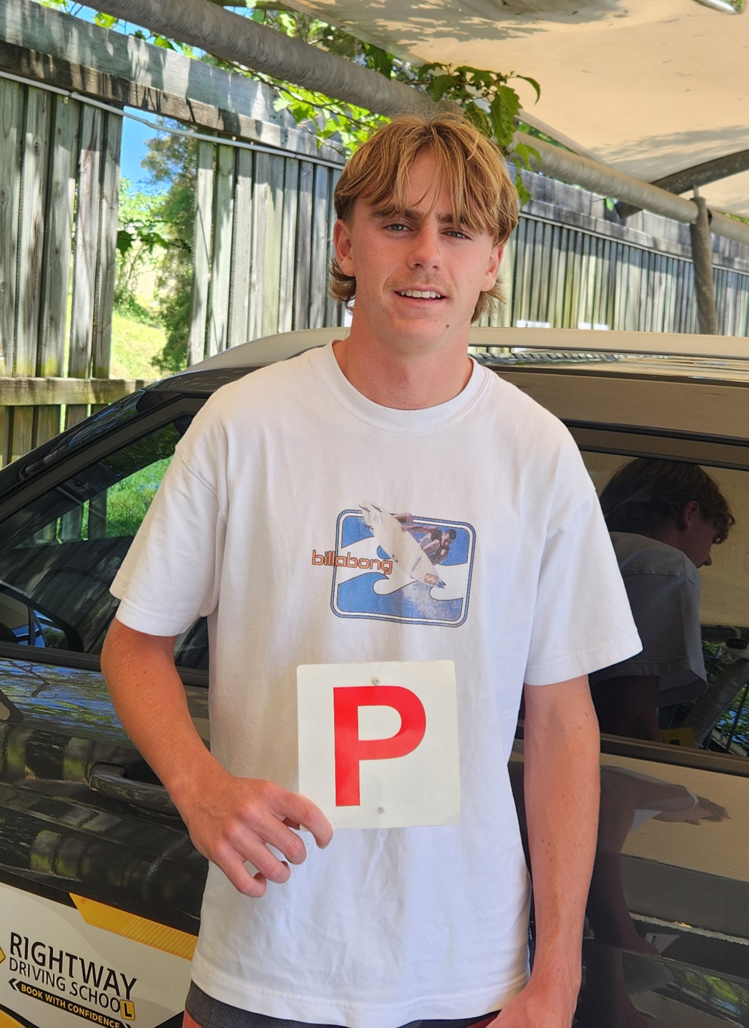 Person in a white printed T-shirt holding a red P plate, with a car and canopy behind.