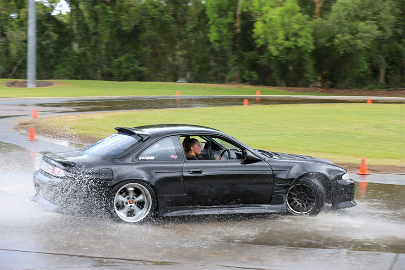 Black car with a femal driver skidding through water on a skid pan.