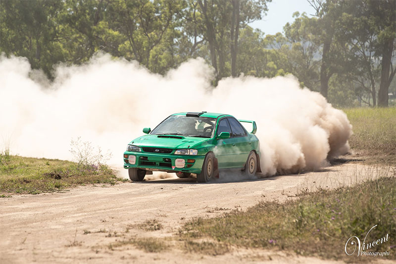 A green rally car driving on a dirt track with lots of dust behind it.