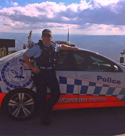 Brad standing at his Highway Patrol car at a lookout with mountain background