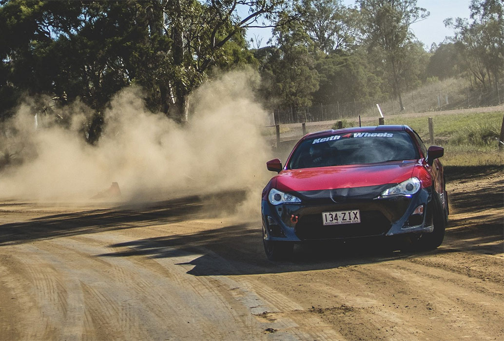 Red car on a dirt rally track with lots of dust behind it.
