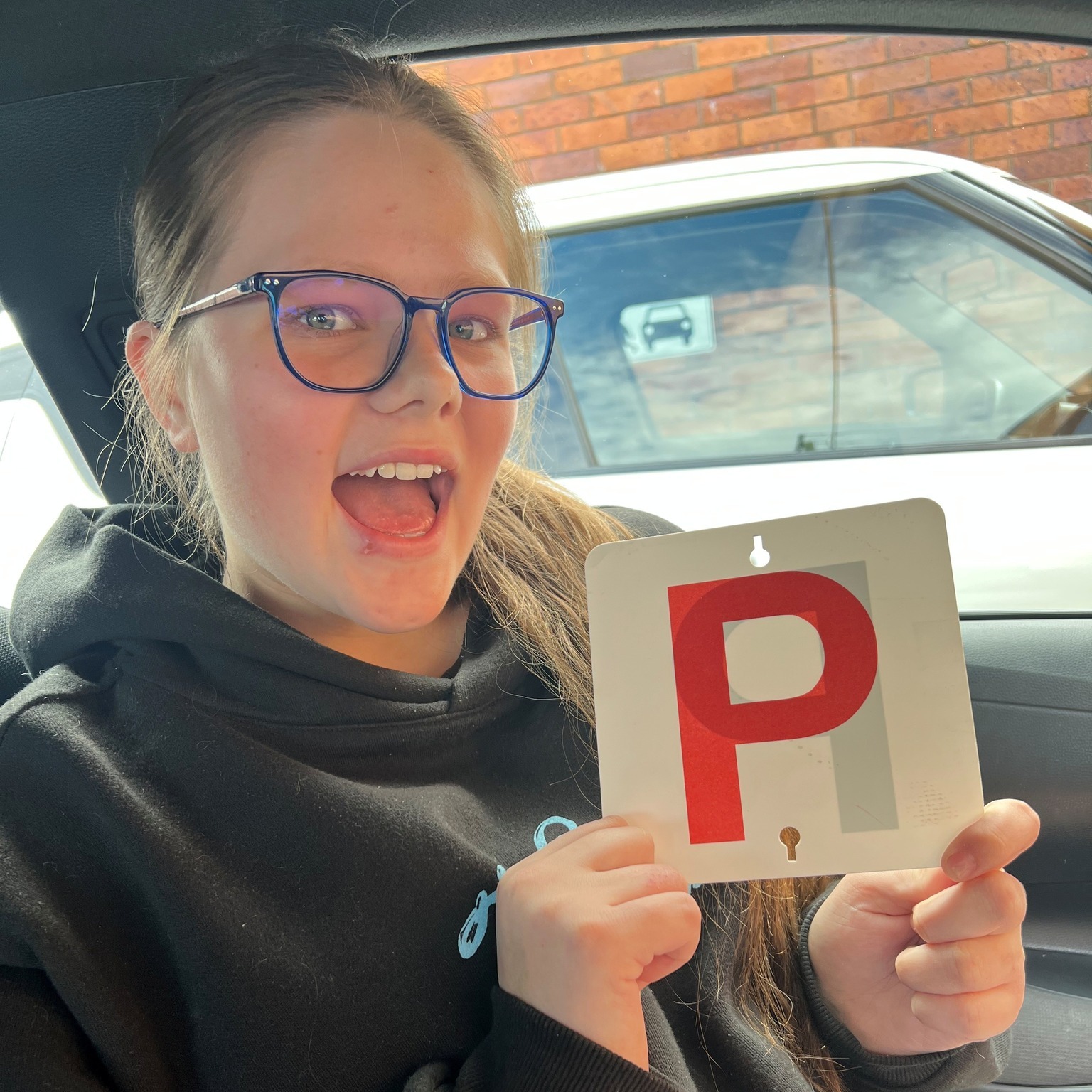 Person wearing glasses seated in a car and holding a red P plate.