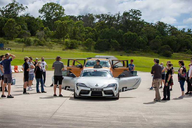 Group of people mingling around cars on a track.