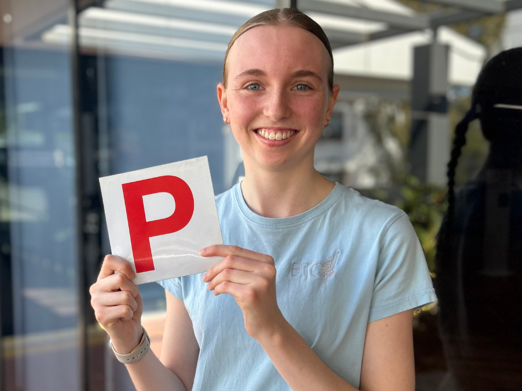 Person in a light-blue top holding a red P plate outside a building