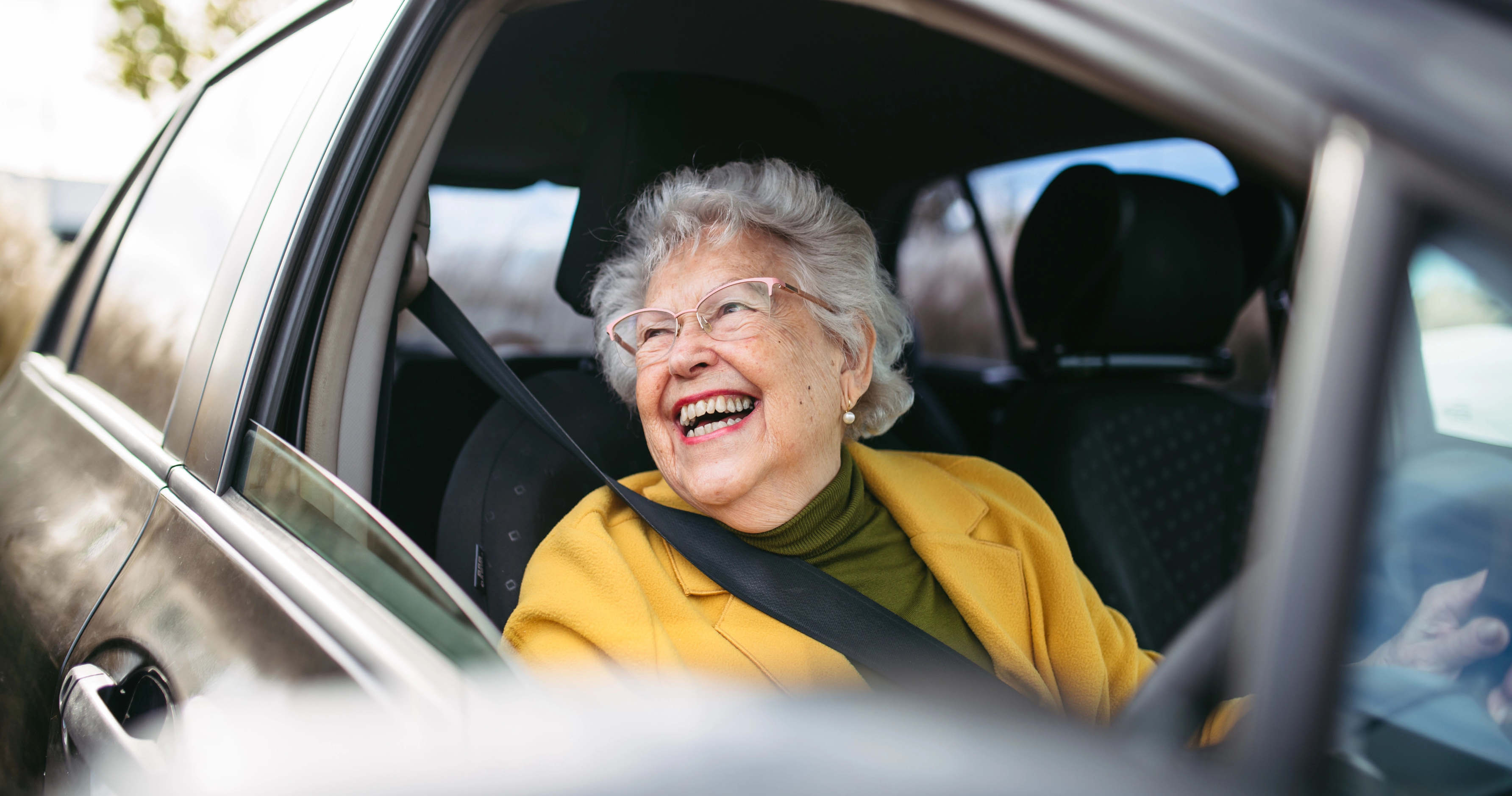 An older woman sits in the driving seat of a car and looks joyously out of the window.