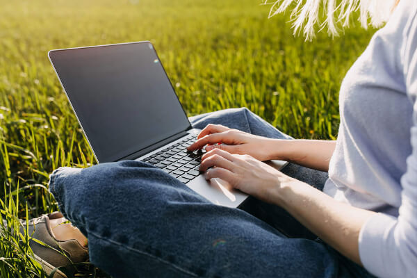 A woman sits in a grassy fiels and uses her laptop.