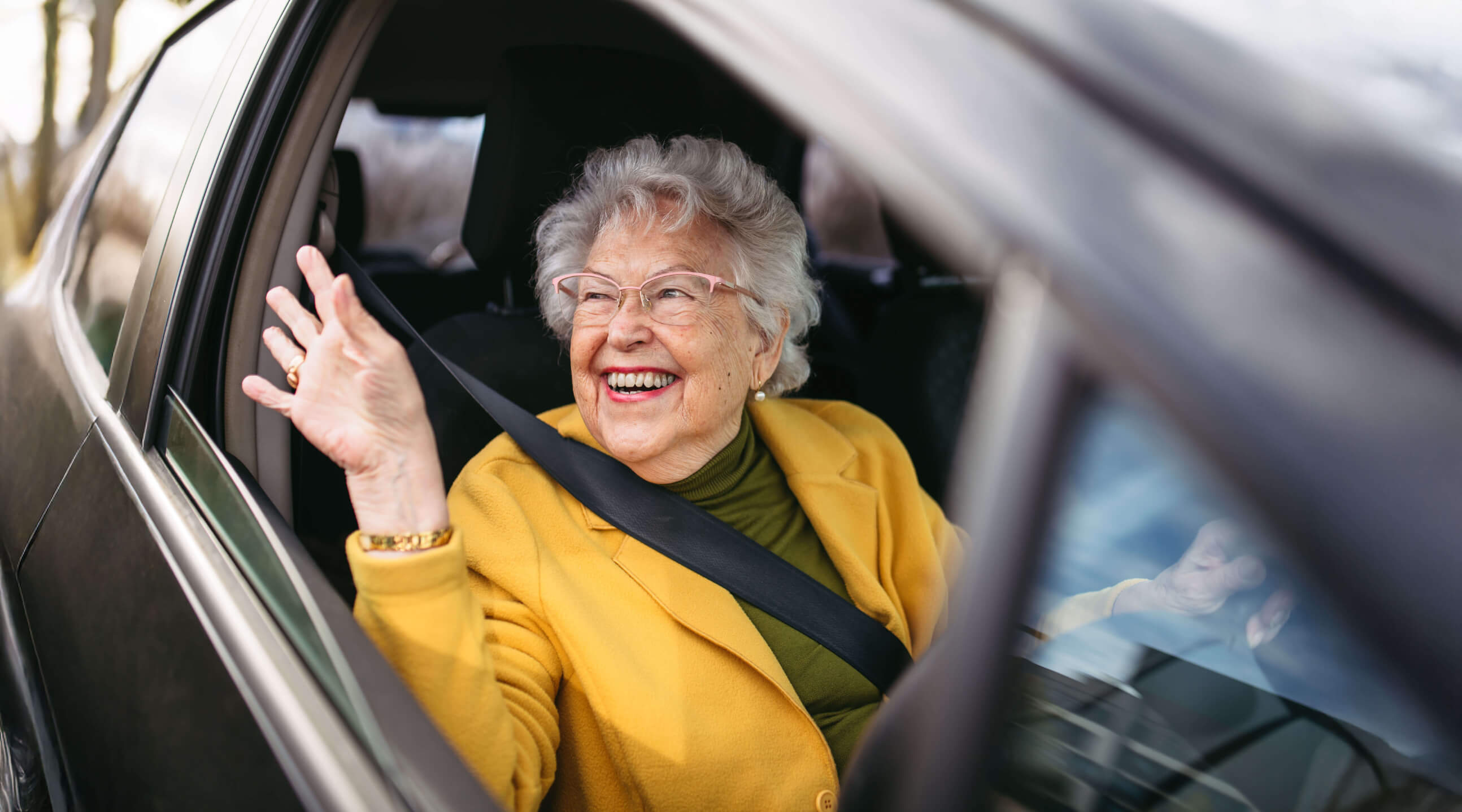 A happy older woman sits in the driving seat of a car and waves out of the window.