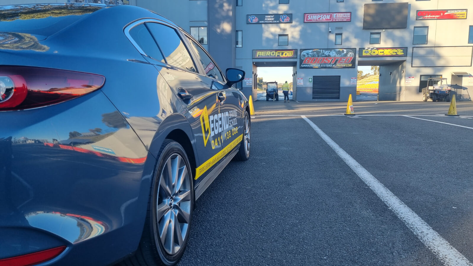 Young smiling man stands next to a driving school car, holding a P plate and displaying a thumbs up gesture.