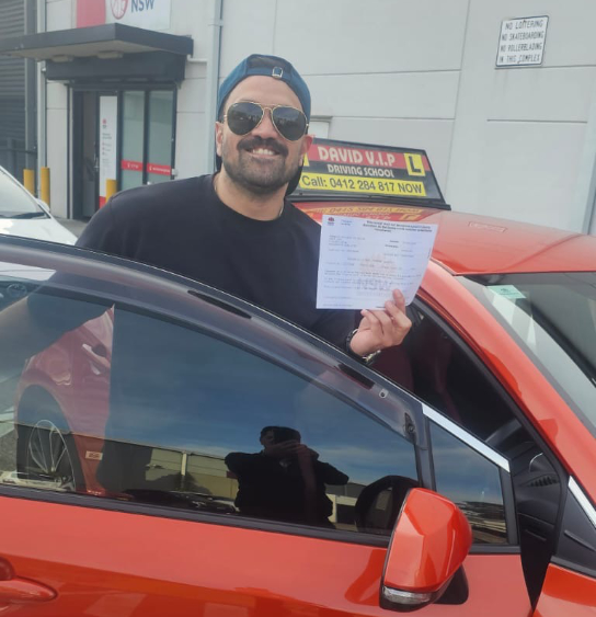 Happy student in sun glasses stands in front of a red Driving Lesson car after passing his Driving Test in Parramatta.