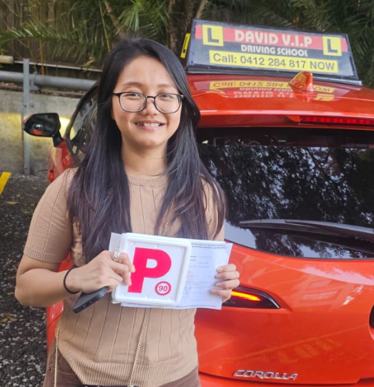 Happy student with glasses stands in front of a red Driving Lesson car after passing her Driving Test in Parramatta.