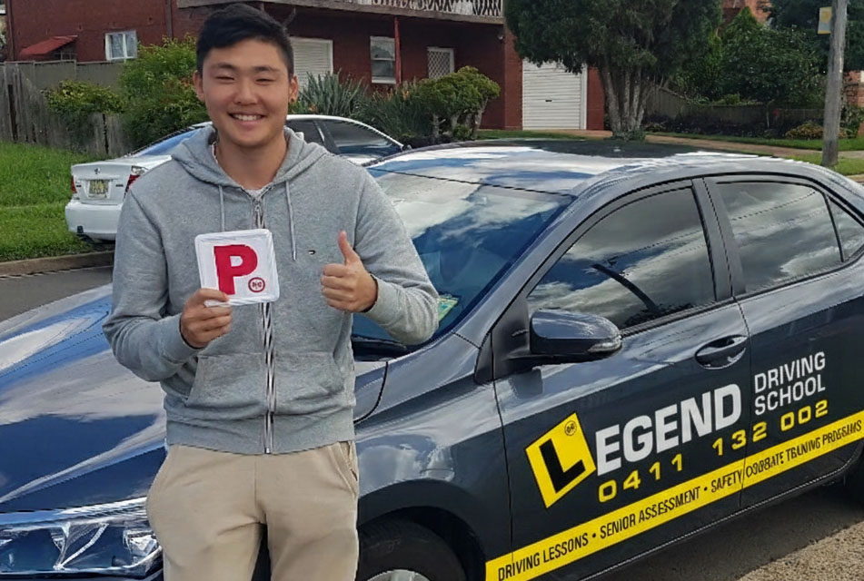 Young smiling man stands next to a driving school car, holding a P plate and displaying a thumbs up gesture.