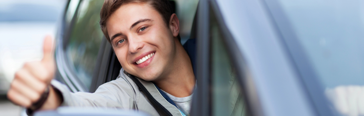 Smiling young man in the driving seat of a car on a driving lesson, while displaying a thumbs up.