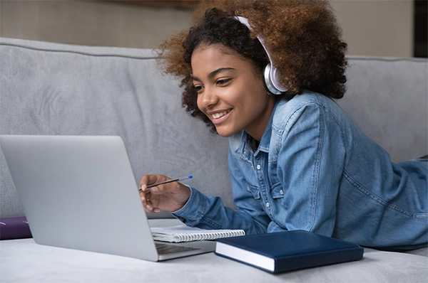 Teen student lying on a sofa with headphones on, smiling while completing an online driver education course.