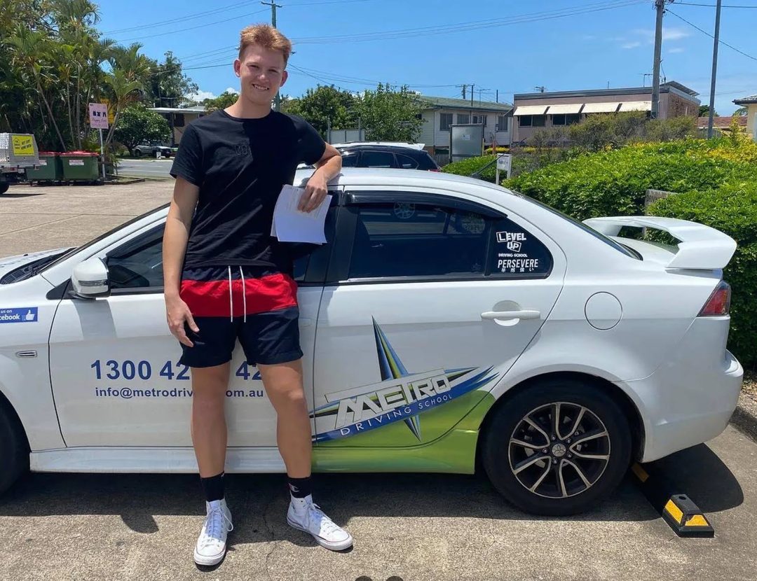 A Metro Driving School learner holding their green P-plates beside a Metro dual-control car after passing the driving test