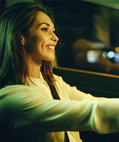 Learner smiling at the wheel of an automatic car while driving after dark, with city lights blurred through the window