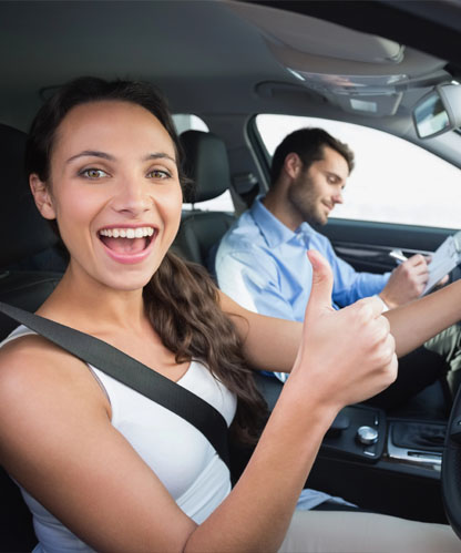 Delighted learner giving a thumbs up in the driver's seat on test day while the instructor beside her completes the paperwork