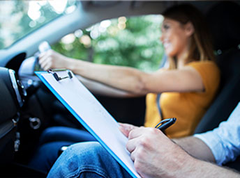 Driving instructor marking an assessment sheet on a clipboard beside a learner at the wheel
