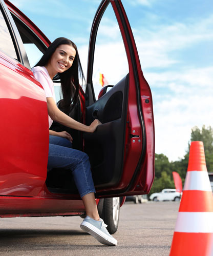 Smiling learner stepping out of the driver's seat of a red automatic car beside a traffic cone at a driving test centre