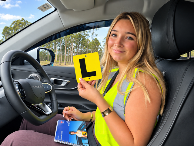 A young woman sitting in the driver's seat of a car, wearing a high-visibility vest and holding up a yellow Australian Learner driver L plate.