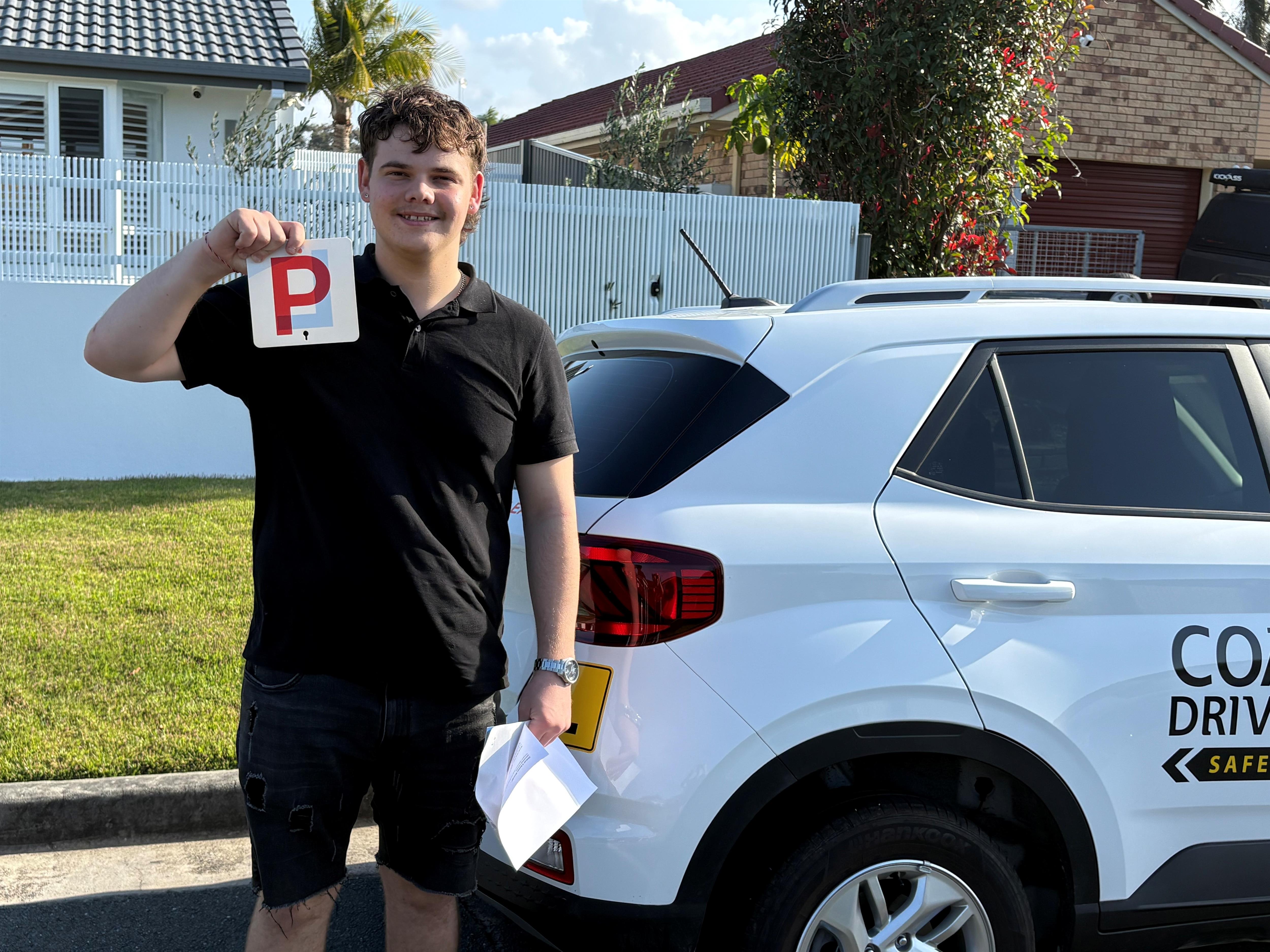 Person in a black polo holding a red P plate beside a white Coastwide car