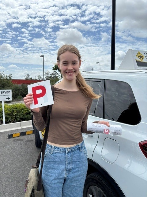 Person in a brown top holding a red P plate beside a white learner car