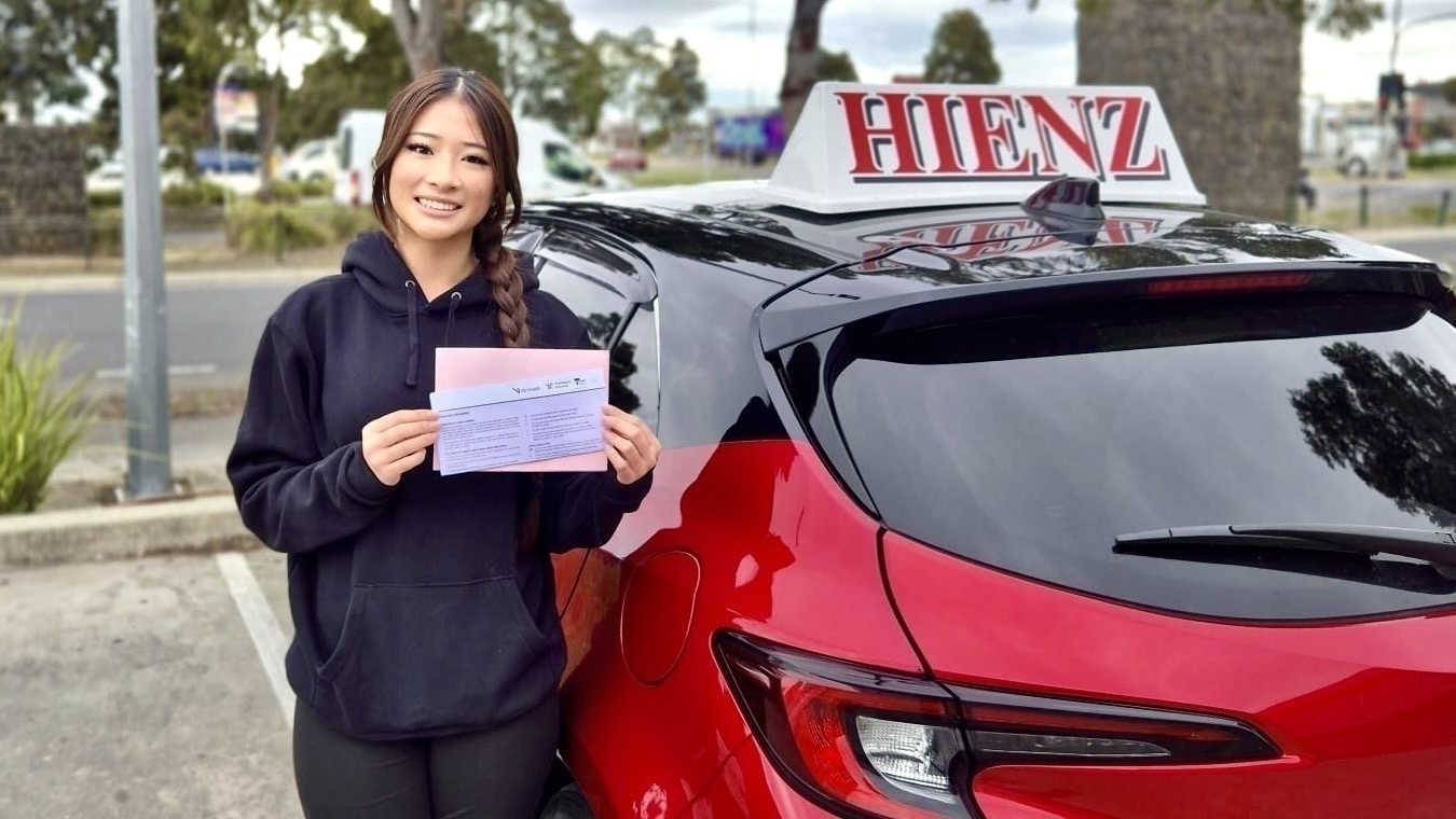 Successful HIENZ student holding paperwork beside a red driving school car.